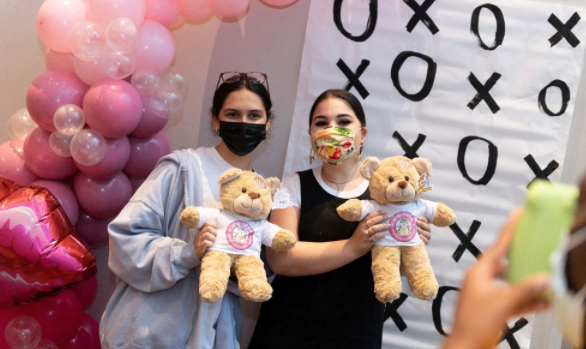 Two friends are holding bears in front of a Valentine balloon arch.